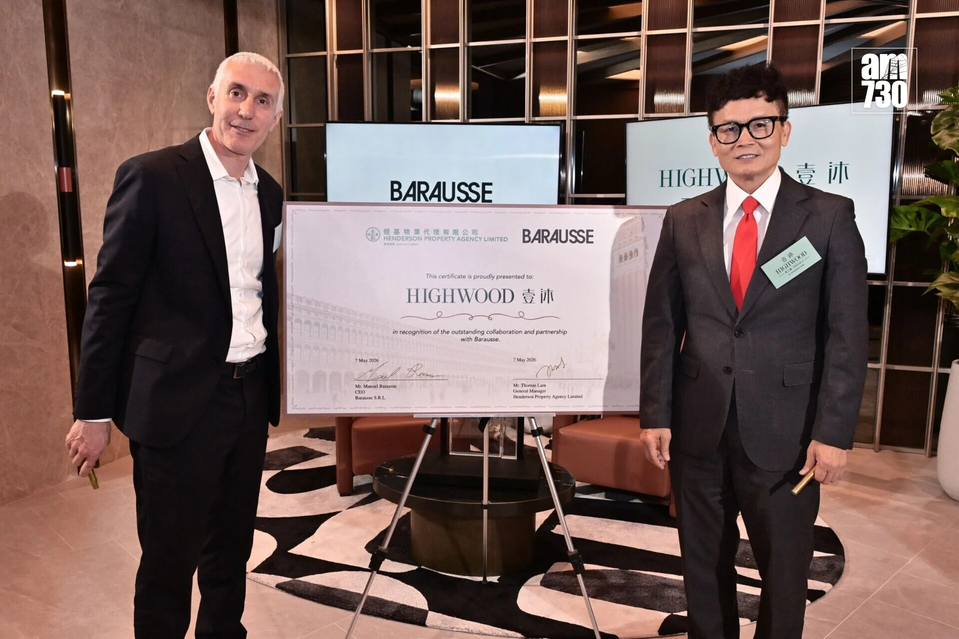 Two men in suits pose with a large certificate labeled HIGHWOOD in a modern lobby setting.
