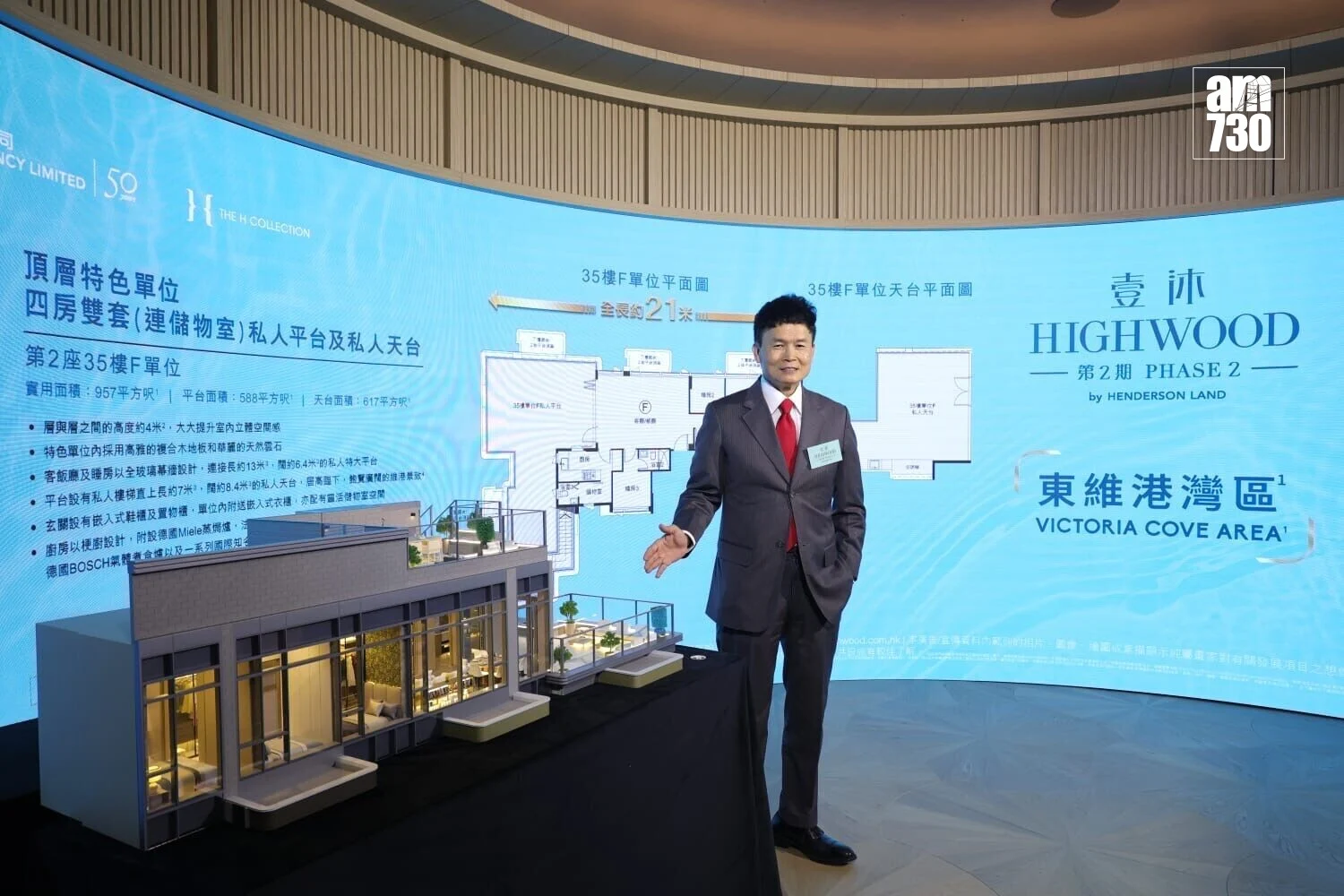 Man in a suit presenting a detailed architectural model of a building at a real estate launch with a blue display backdrop