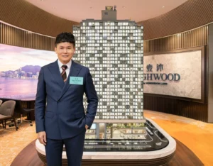 Smiling man in a navy suit stands beside a tall apartment building model in a showroom with a HIGHWOOD sign on the wall behind him