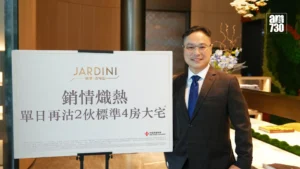 Man in a brown suit with a blue tie posing beside a large white sign displaying the JARDIN logo and Chinese text