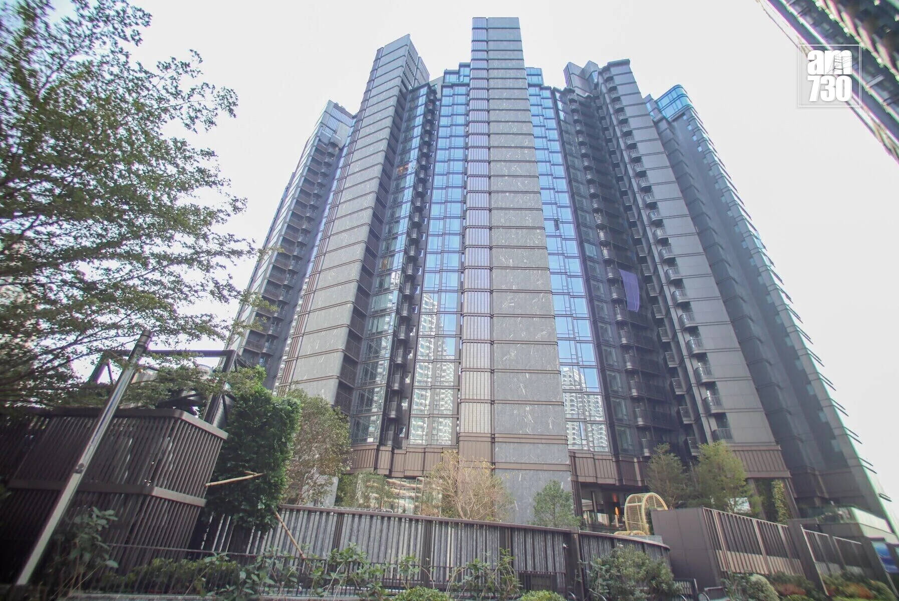 Tall modern skyscraper photographed from the ground looking upward, with trees on the left and a fence in the foreground.
