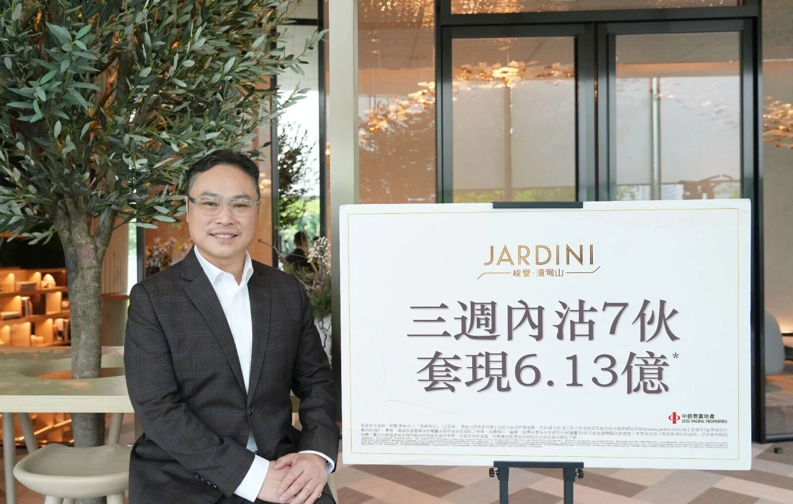 Man in a dark suit smiles beside a large white sign with the JARDINI logo and Chinese text, in a bright lobby with plants behind him.