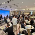 Crowded business networking event in a modern hall people sit at round tables chat and review materials near a large blue presentation screen and buffet area