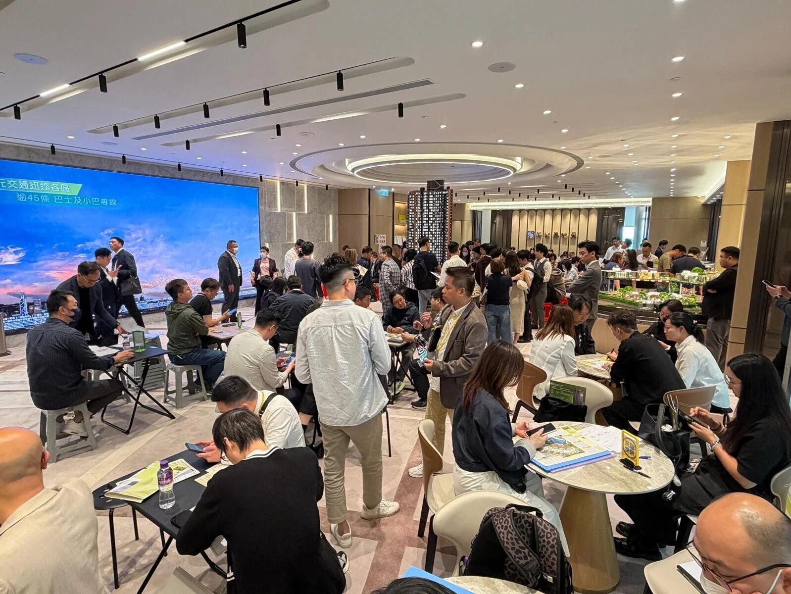 Crowded business networking event in a modern hall people sit at round tables chat and review materials near a large blue presentation screen and buffet area