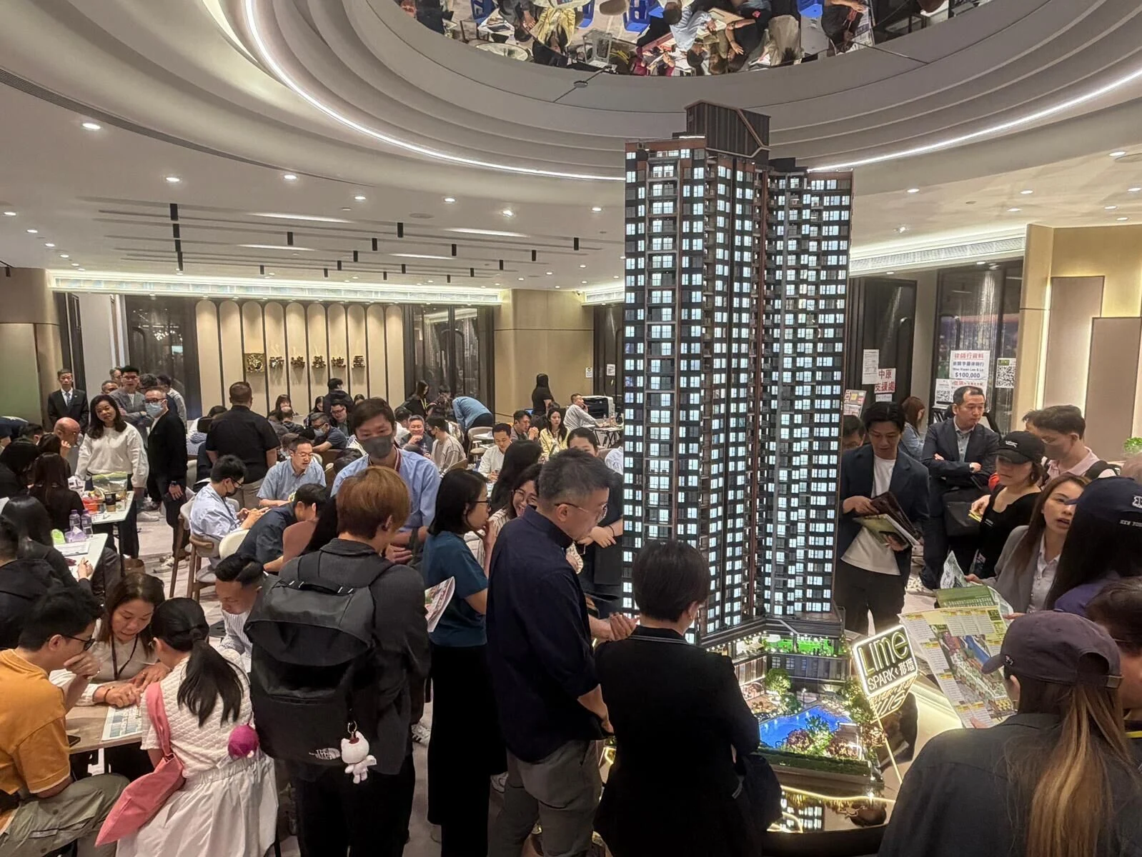 Crowded real estate sales showroom around a large scale-model of a tall apartment building, with people examining brochures and talking with agents.
