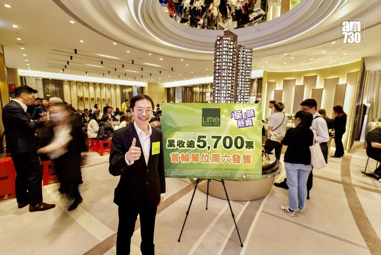 Smiling man in a suit gives a thumbs-up beside a large green sign announcing 5,700 units sold at an indoor real estate event. Crowds are gathered in the background.