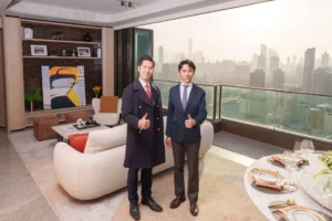 Two professionally dressed men stand in a modern living room with a city skyline view giving thumbs up to the camera