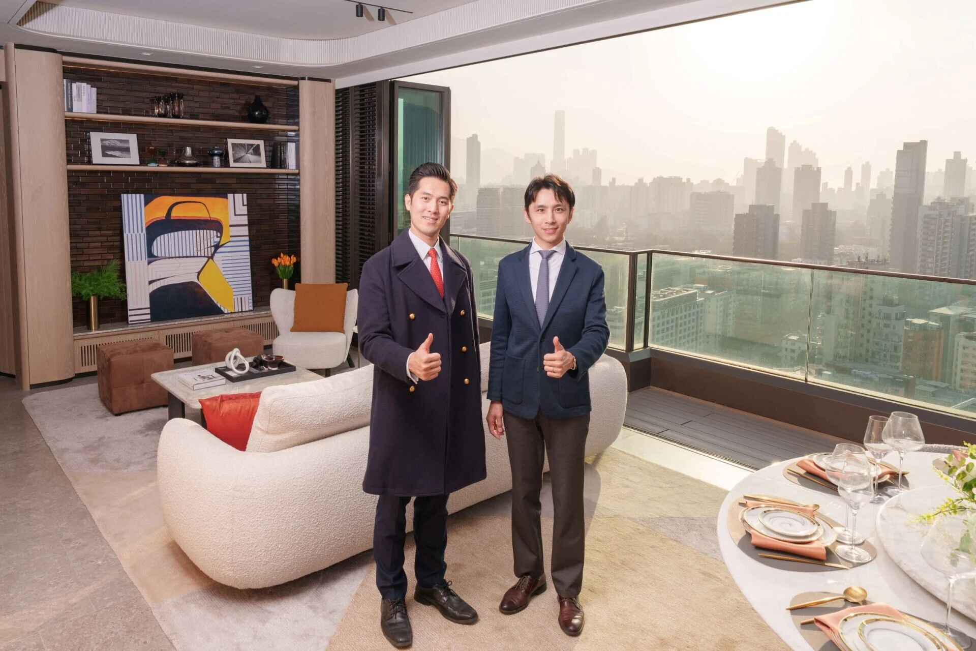 Two professionally dressed men stand in a modern living room with a city skyline view giving thumbs up to the camera