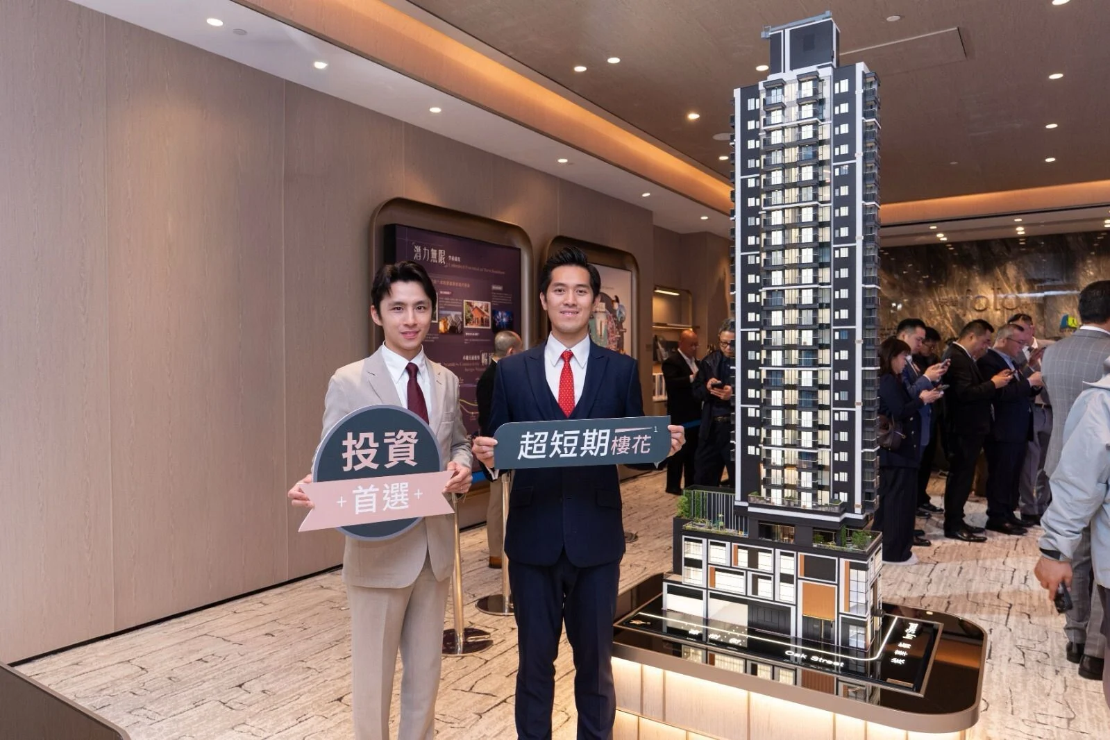 Two men in suits pose at a real estate showroom, holding Chinese signs beside a tall architectural tower model.