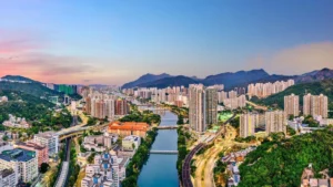 City skyline along a river with green hills and distant mountains at sunset