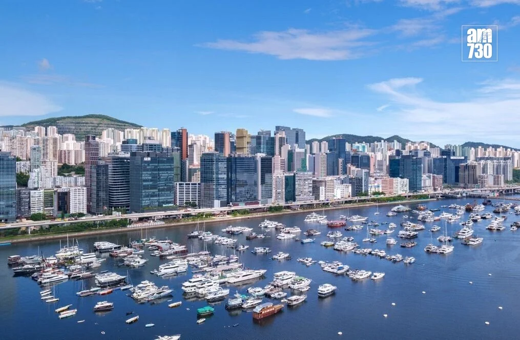 Harbor filled with moored yachts and speedboats, with a dense city skyline along the waterfront under a blue sky.