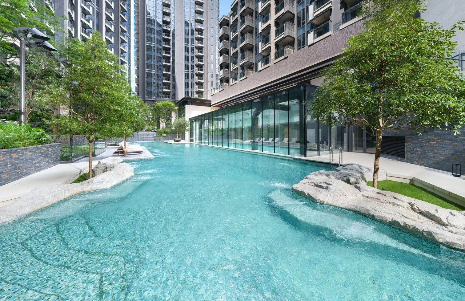 Outdoor pool area at a high rise residential complex with glass walled building and turquoise water surrounded by trees and stone features