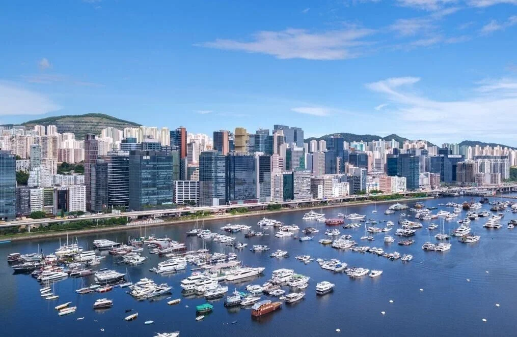 A coastal city skyline with tall modern buildings lined along a waterway filled with numerous moored yachts and boats.