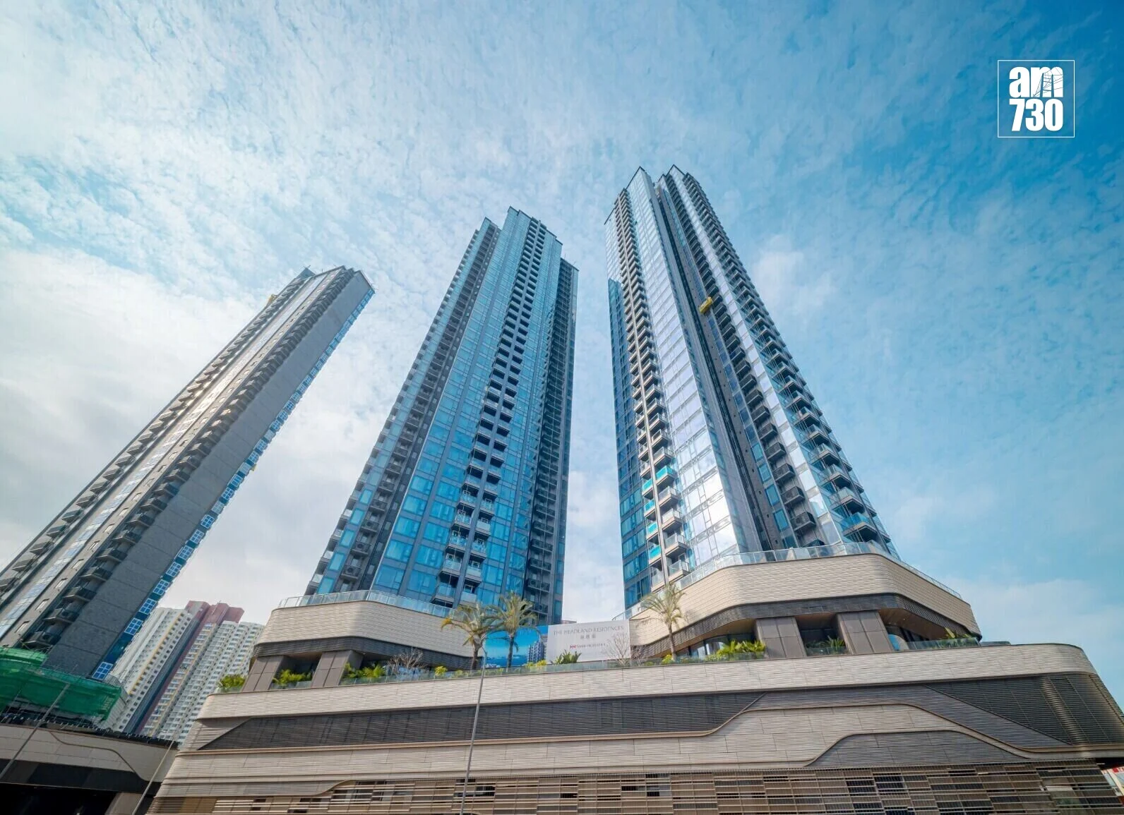 Three glass high-rise towers rise into a blue sky, with a curved base and palm trees at the entrance; am730 logo in the corner.