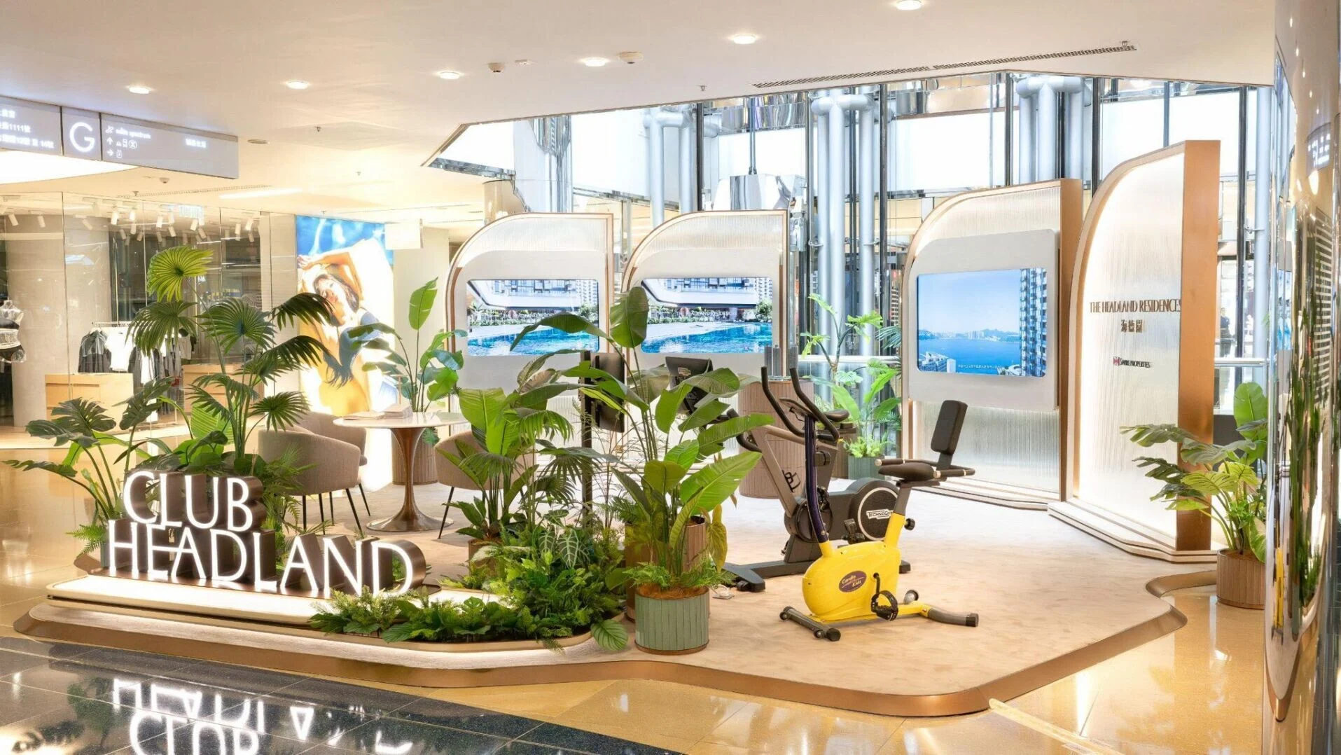 Showroom interior with exercise bikes and lush potted plants a glowing CLUB HEADLAND sign and curved display panels behind glass walls