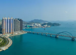 City skyline with tall high rise buildings along a blue harbor and a curved bridge stretching across the water hills in the distance