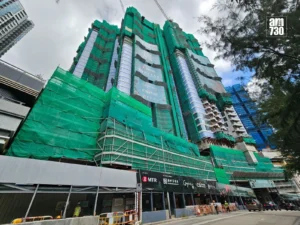 Tall high rise under construction wrapped in green safety netting with scaffolding and cranes above along a city street with barriers