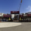 Entrance to a construction site with a tall crane and red Pavilia Rosa banners visible against a blue sky