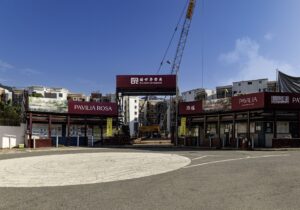 Entrance to a construction site with a tall crane and red Pavilia Rosa banners visible against a blue sky