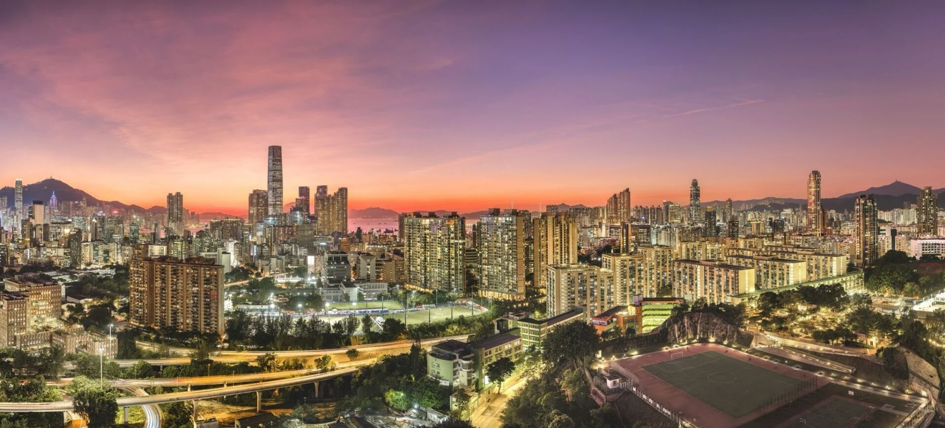 Panoramic city skyline at sunset with an orange to purple sky, dense high-rise buildings, and a foreground sports field.