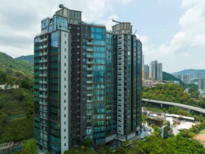 Tall glass fronted apartment building rising beside green hills under a blue sky with scattered clouds