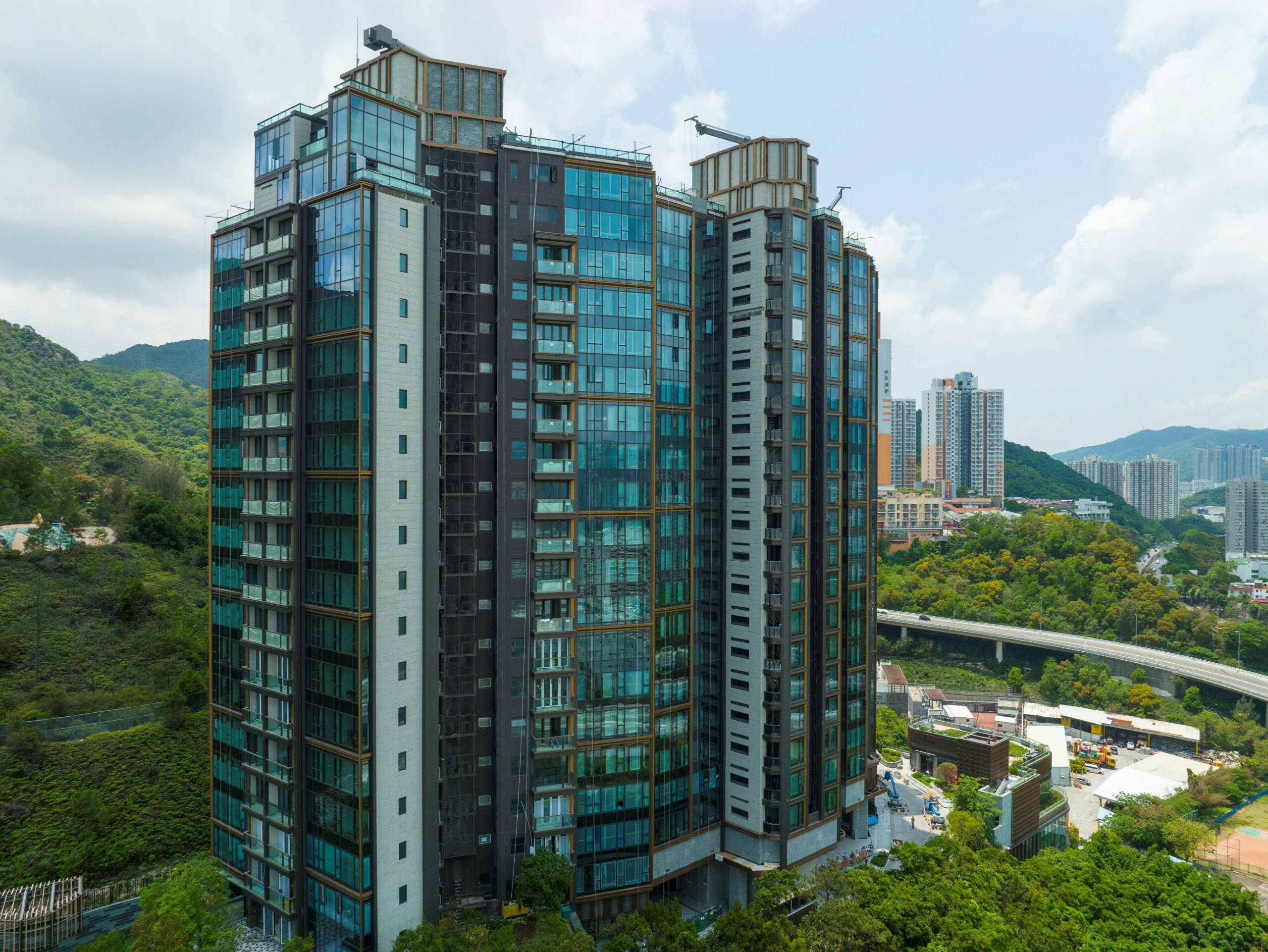 Tall glass-fronted apartment building rising beside green hills under a blue sky with scattered clouds.