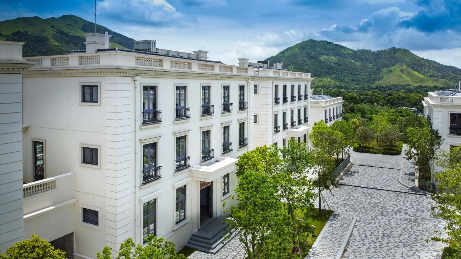 White multi-story residential building with balconies, trees, and a cobblestone courtyard set against green hills behind it.