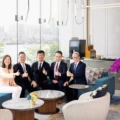 Five professionals in business attire sit on a blue sectional in a bright modern lounge giving thumbs up with a city skyline visible through glass walls and purple orchids on the right