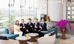 Five professionals in business attire sit on a blue sectional in a bright modern lounge giving thumbs up with a city skyline visible through glass walls and purple orchids on the right