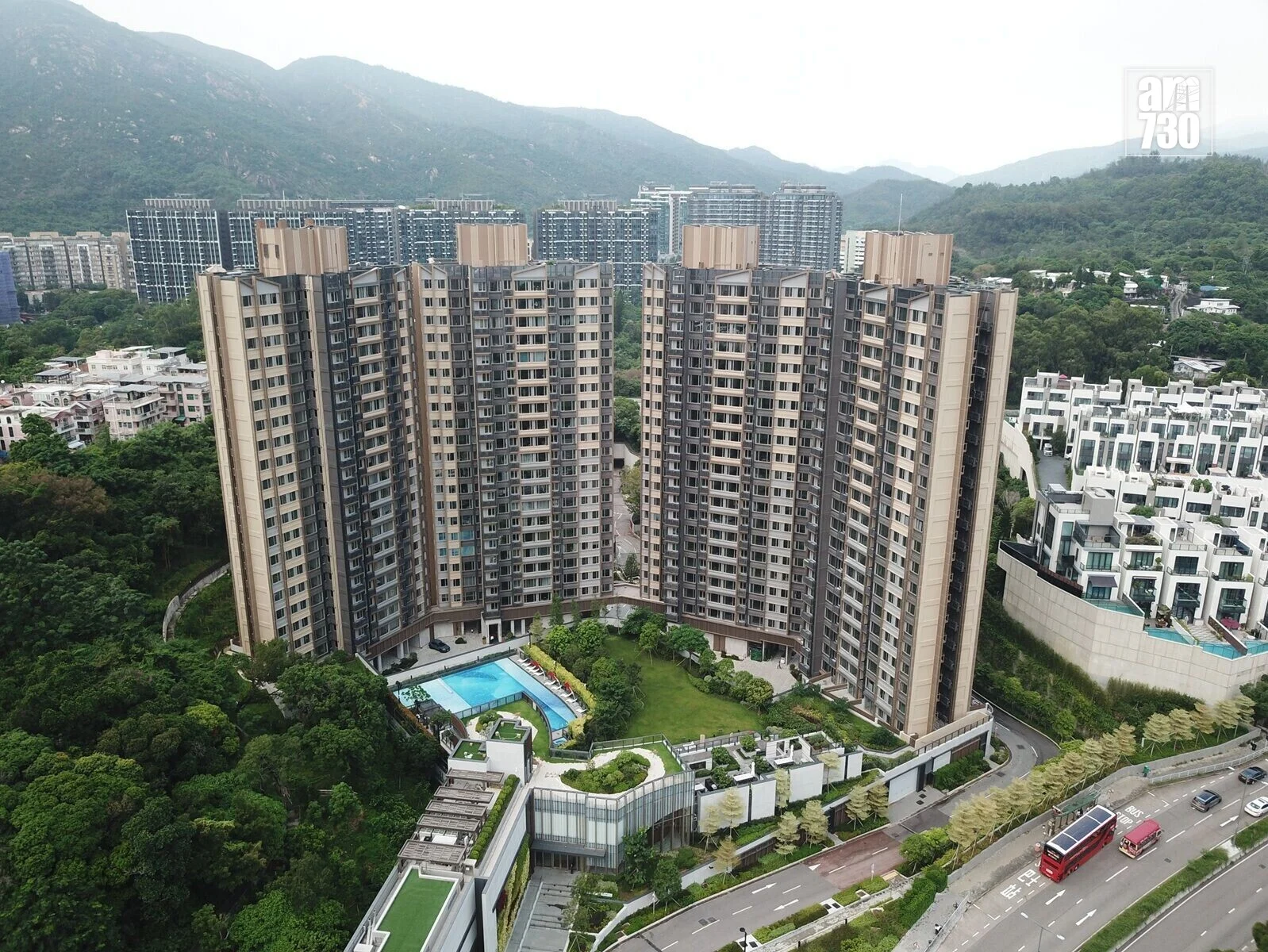 Aerial view of a large beige apartment complex with curved towers, surrounded by greenery and a swimming pool area at the center, mountains in the distance.