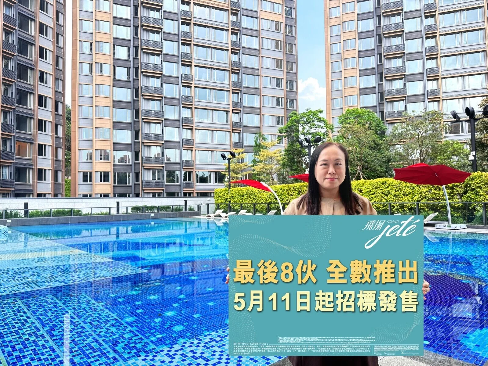 Woman stands at a blue tiled poolside holding a teal promotional sign with Chinese text, tall apartment buildings in the background and red umbrellas nearby.