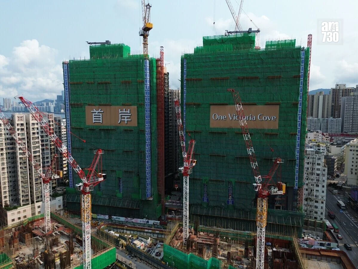 Two under-construction high-rise buildings covered in green netting with cranes nearby; sign reads 'One Victoria Cove' on the right tower.