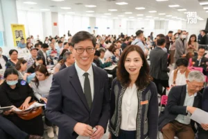 Two smiling colleagues pose for a photo at a crowded conference hall with attendees seated and mingling in the background