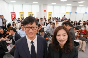 Man in a dark suit and glasses taking a selfie with a smiling woman beside him at a crowded conference hall