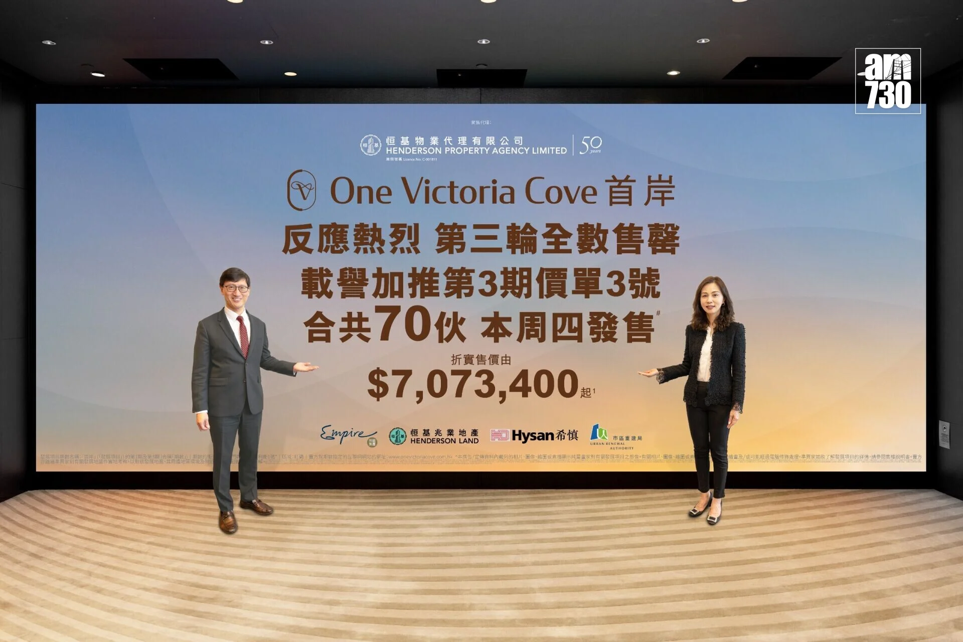 Two presenters in business attire stand on a stage beside a large gradient backdrop reading 'One Victoria Cove' with Chinese text and logos.