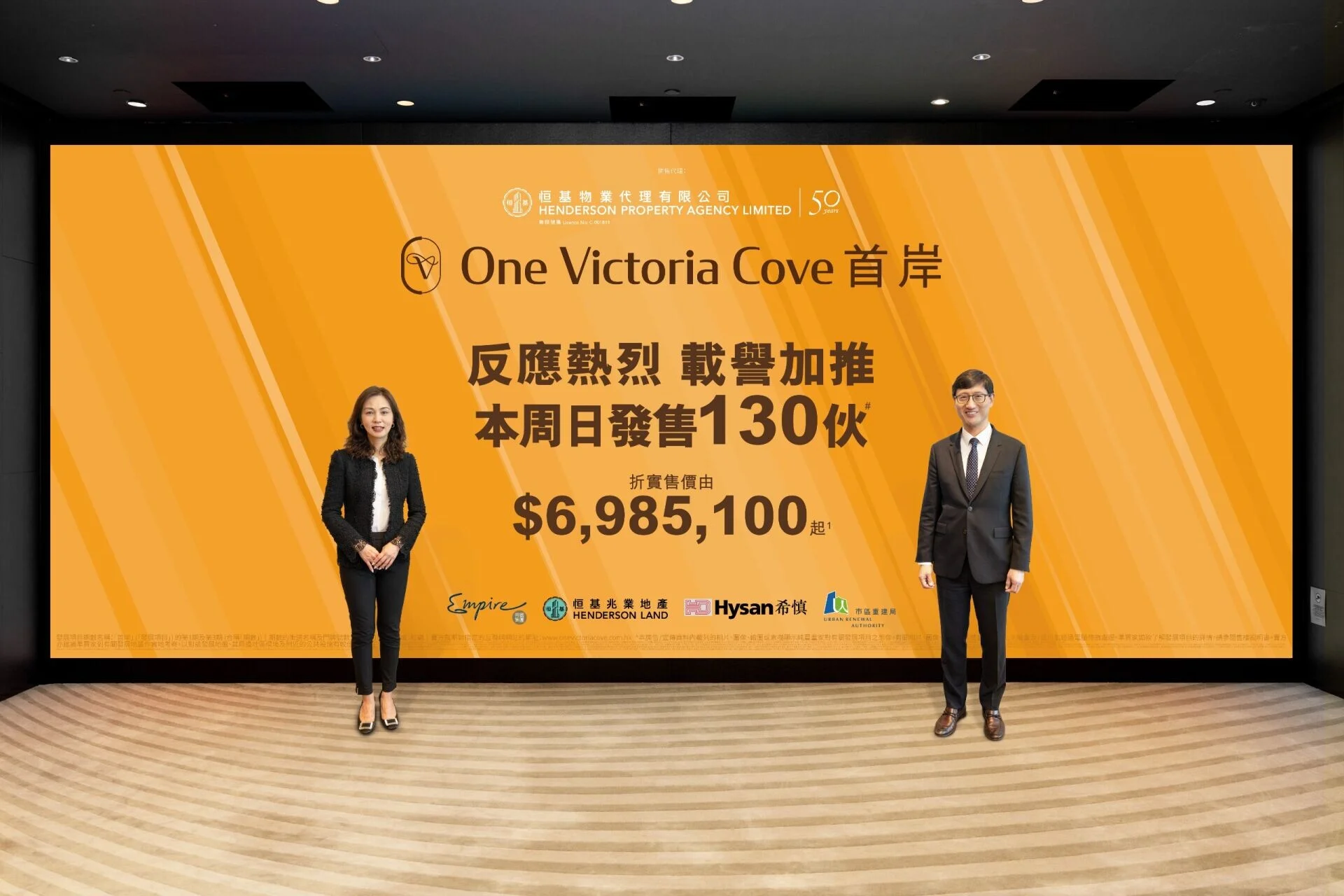 Two professionally dressed individuals stand beside an orange promotional backdrop for 'One Victoria Cove,' featuring Chinese text and pricing details.
