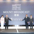 Seven professionals in formal attire stand on a stage in front of a blue backdrop reading MOUNT BROADCAST with company logos posing for a photo